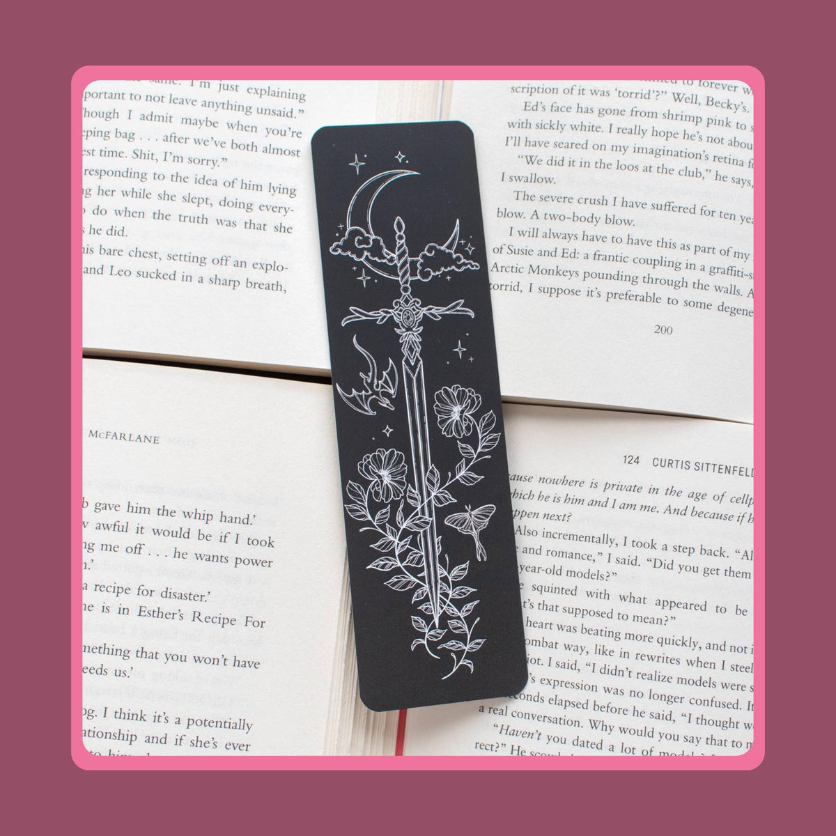 May include: A rectangular black bookmark featuring a white line art illustration of a sword pointing downwards, surrounded by a crescent moon, clouds, stars, a small dragon, a moth, and floral vines, resting on top of open book pages with printed text.