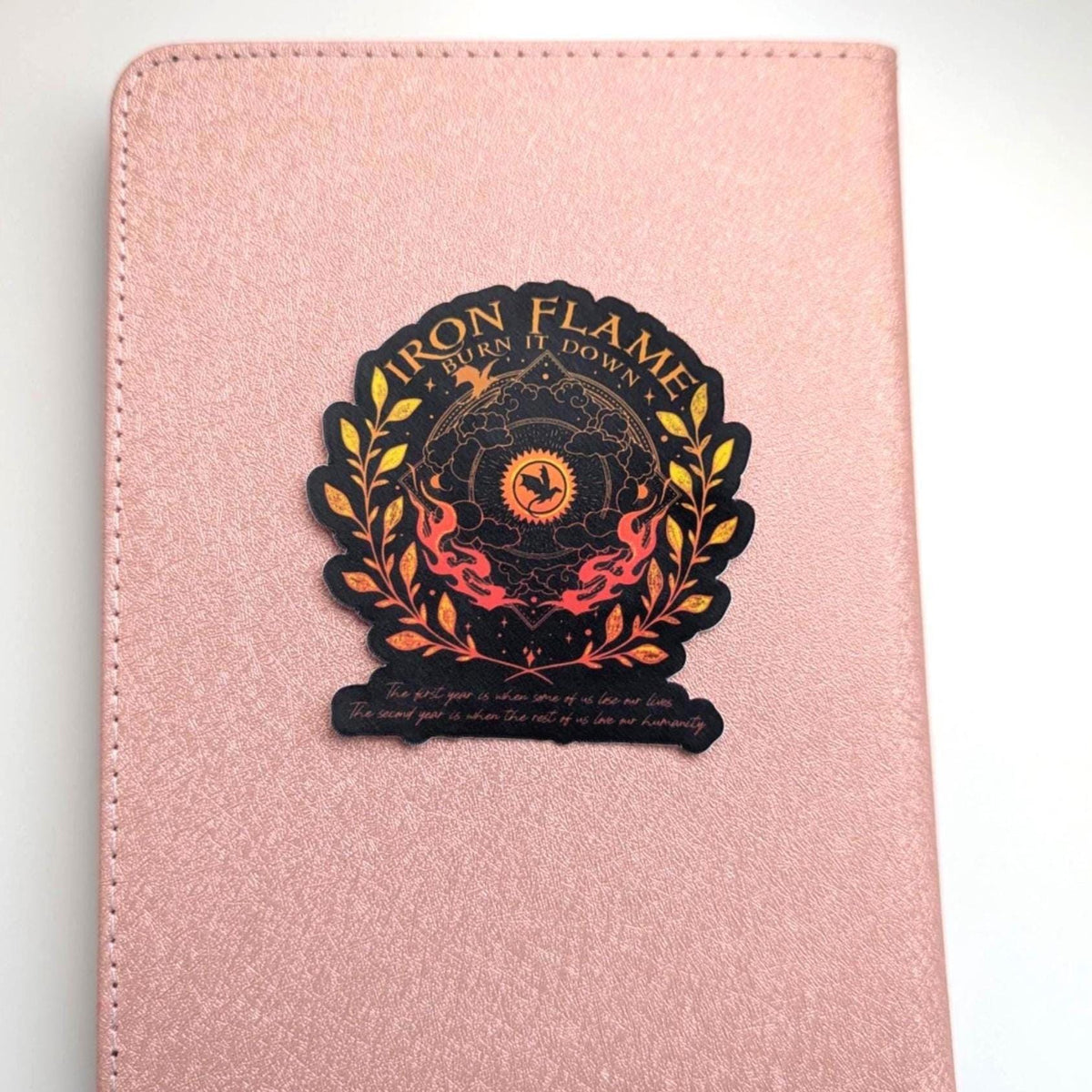 May include: A rose gold textured notebook featuring a centred sticker with the text Iron Flame Burn It Down. The sticker displays a black and orange circular emblem with a dragon silhouette, surrounded by golden laurel leaves and flames.