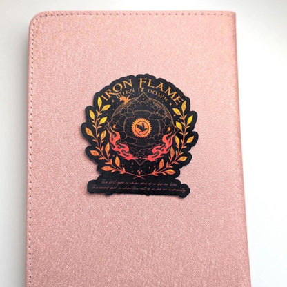 May include: A rose gold textured notebook featuring a centred sticker with the text Iron Flame Burn It Down. The sticker displays a black and orange circular emblem with a dragon silhouette, surrounded by golden laurel leaves and flames.