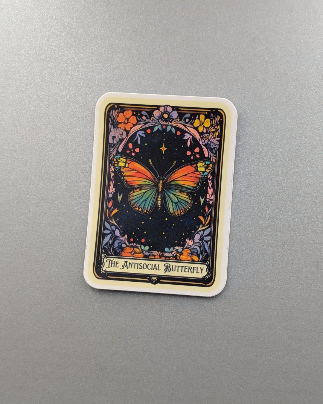 Angie's Creations waterproof die-cut sticker featuring a tarot card style design titled The Antisocial Butterfly, with a vibrant rainbow butterfly centred on a dark floral botanical background with gold star accents and ornate card border. Bookish humor and dark academia inspired waterproof sticker designed in Ottawa Canada, perfect for laptops, water bottles, journals, and Kindle cases.