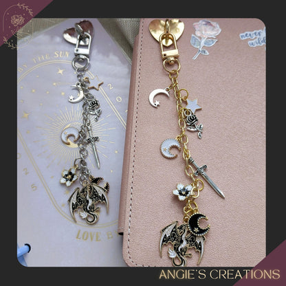 A silver chain Kindle charm displayed on a pastel planner, featuring dragon, moon, sword, star, and rose charms in a romantasy aesthetic.
