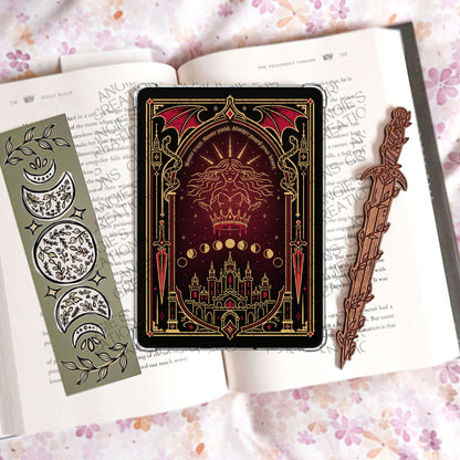 May include: A rectangular tablet case featuring a dark red and black illustration of a figure with a crown, a castle, and the text 'Never trust. Never yield. Always guard your heart.' Flanked by a green moon phase bookmark and a wooden sword bookmark.