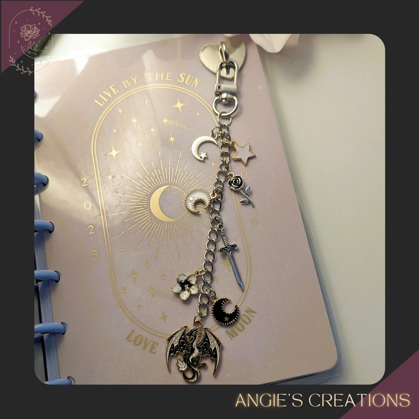 A silver chain Kindle charm displayed on a pastel planner, featuring dragon, moon, sword, star, and rose charms in a romantasy aesthetic.