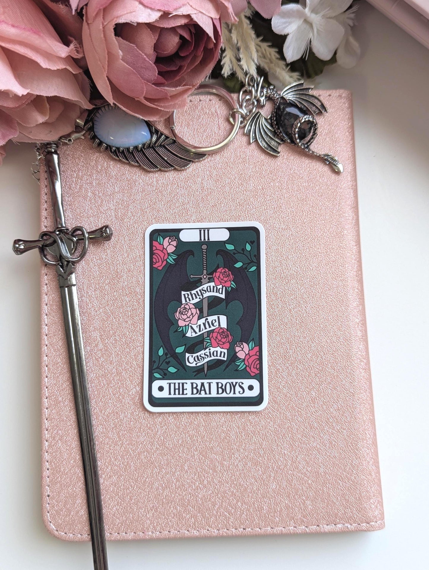 May include: A rose gold notebook featuring a rectangular sticker with a dark green background, a central sword, black wings, and pink roses. The sticker displays the text Rhysand, Azriel, Cassian, and THE BAT BOYS. A silver sword bookmark and dragon charm are attached.