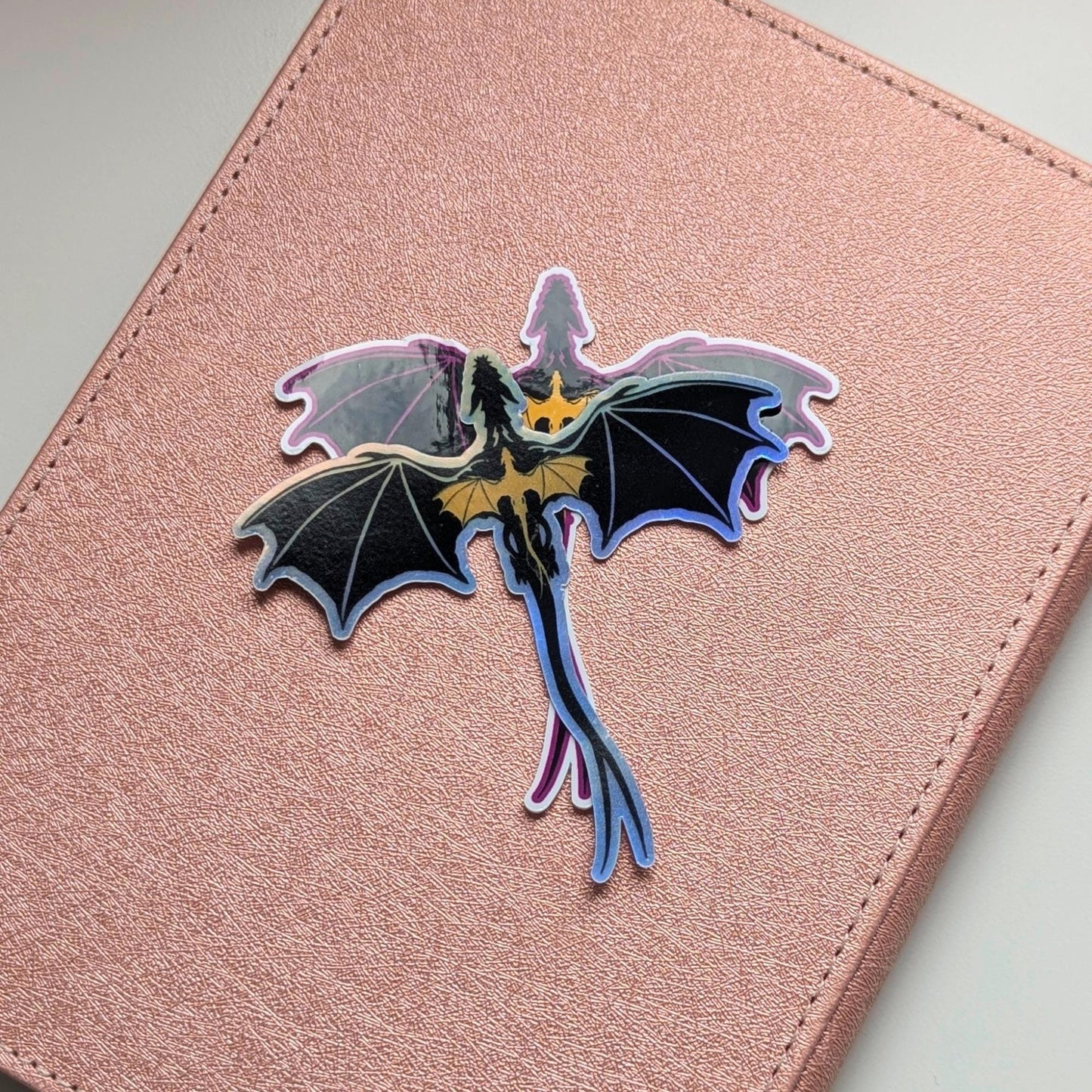 May include: A holographic dragon sticker with black wings and a gold chest, on a pink background. The dragon is depicted in flight, with a long tail. The sticker has an iridescent, shiny finish.