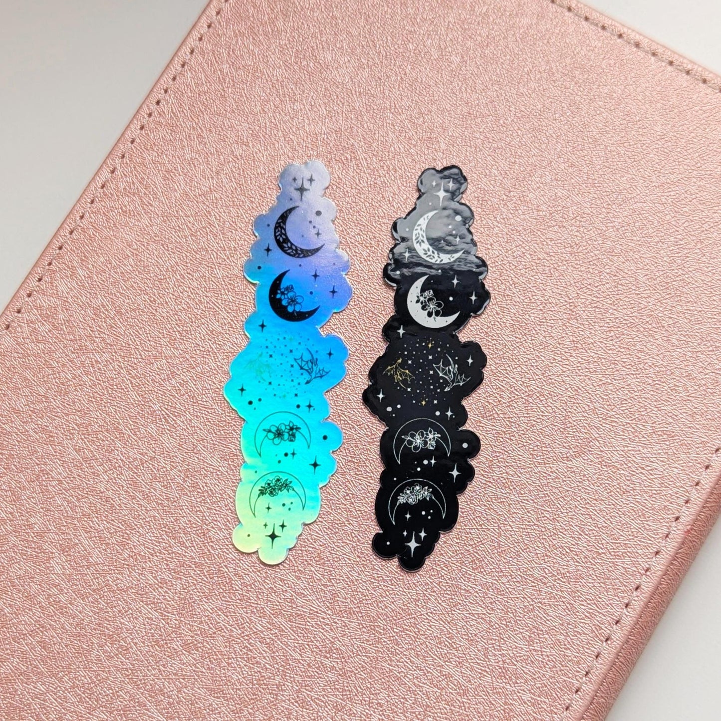 May include: Two holographic stickers with a celestial design of crescent moons, stars, and floral accents. One has a blue and green gradient, the other is black and white. They are placed on a pink surface.