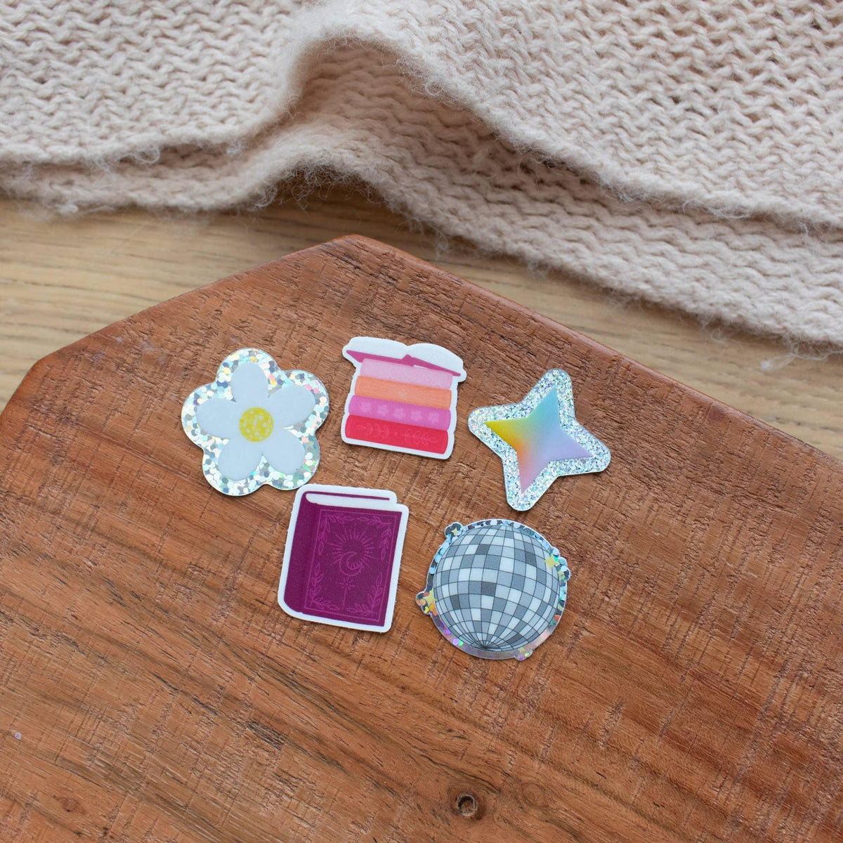 Set of five Y2K-style mini stickers including a holographic daisy, a pink ombre stack, a holographic star, a purple glittery book, and a checkered disco ball, all on wood background.