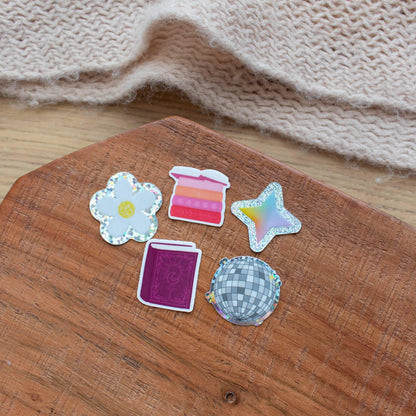 Set of five Y2K-style mini stickers including a holographic daisy, a pink ombre stack, a holographic star, a purple glittery book, and a checkered disco ball, all on wood background.