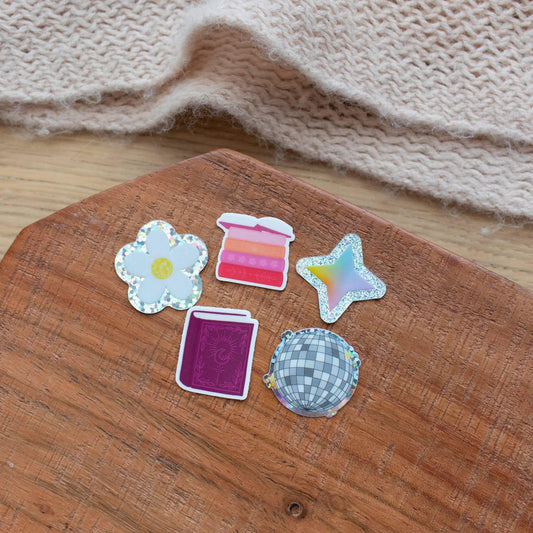 Set of five Y2K-style mini stickers including a holographic daisy, a pink ombre stack, a holographic star, a purple glittery book, and a checkered disco ball, all on wood background.