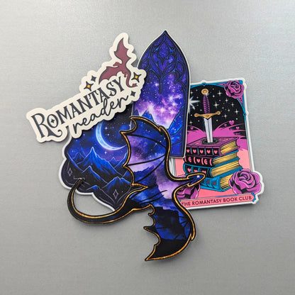 May include: A set of fantasy-themed stickers with a dragon, a starry night scene, and a sword in a book. The stickers are predominantly purple, blue, and pink, with gold detailing. The text "Romantasy reader" is also included.