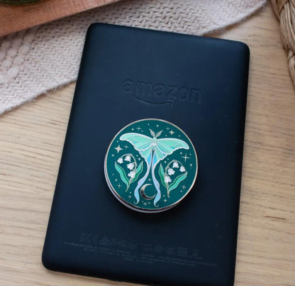 Dark green enamel Kindle grip featuring a pale teal luna moth centered above a crescent moon, flanked by white lily of the valley flowers.