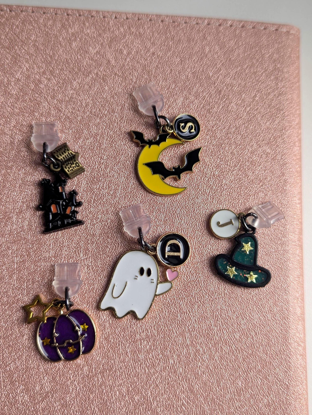 May include: Five metal charms with clear plastic backings. One charm is a black haunted house with a book, one is a yellow crescent moon with black bats and a letter S, one is a white ghost with a letter D, one is a green witch hat with a letter J, and one is a purple pumpkin with gold stars.