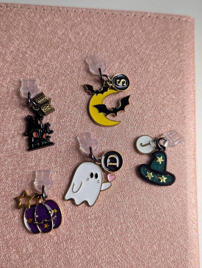 May include: Five metal charms with clear plastic backings. One charm is a black haunted house with a book, one is a yellow crescent moon with black bats and a letter S, one is a white ghost with a letter D, one is a green witch hat with a letter J, and one is a purple pumpkin with gold stars.