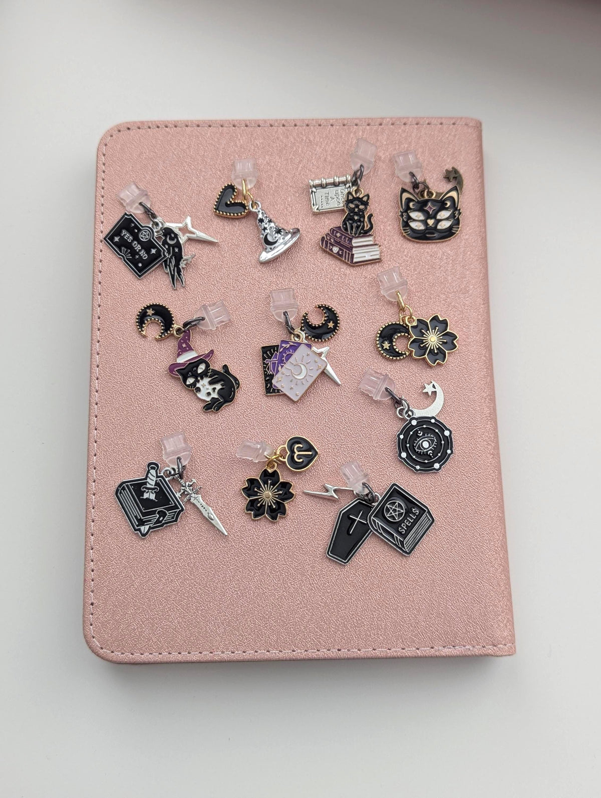 May include: A collection of eleven metal enamel charms featuring various designs including a cat, a witch hat, a coffin, a book labelled SPELLS, a heart, a flower, and a crescent moon, all attached to clear plastic clips on a textured pink surface.