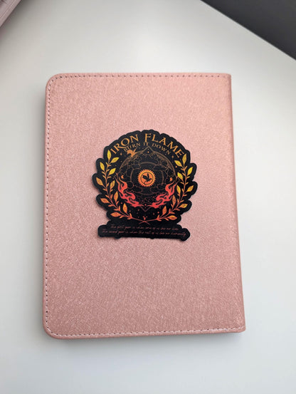 May include: A rose gold textured notebook featuring a centred sticker with the text Iron Flame Burn It Down. The sticker displays a black and orange circular emblem with a dragon silhouette, surrounded by golden laurel leaves and flames.