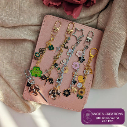 May include: Several handcrafted keychains with charms such as a four-leaf clover, a star, flowers, and a dragon. The keychains are displayed on a pink notebook. The image includes the text "ANGIE'S CREATIONS gifts hand-crafted with love."