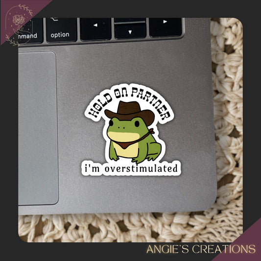 a sticker with a cartoon frog wearing a cowboy hat and the text "HOLD ON PARTNER! I'M OVERSTIMULATED" on a laptop keyboard.