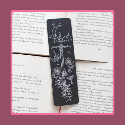 May include: A rectangular black bookmark featuring a white line art illustration of a sword pointing downwards, surrounded by a crescent moon, clouds, stars, a small dragon, a moth, and floral vines, resting on top of open book pages with printed text.