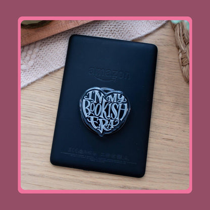 Black heart-shaped acrylic charm with white "In My Bookish Era" script lettering, sold as a matching Kindle grip and phone dust plug set.