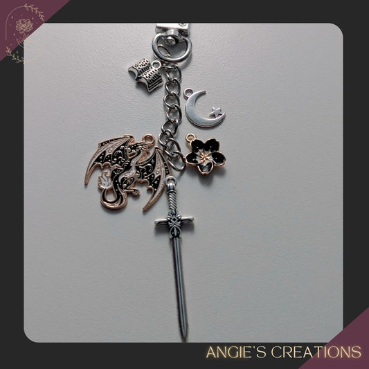 A handmade romantasy-themed Kindle charm chain featuring a detailed dragon charm, silver sword, crescent moon, flower charm, and tiny open book, attached to a silver lobster clasp. Perfect for fantasy readers and bookish accessory lovers.