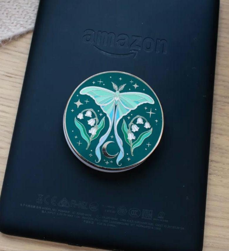 Dark green enamel Kindle grip featuring a pale teal luna moth centered above a crescent moon, flanked by white lily of the valley flowers.