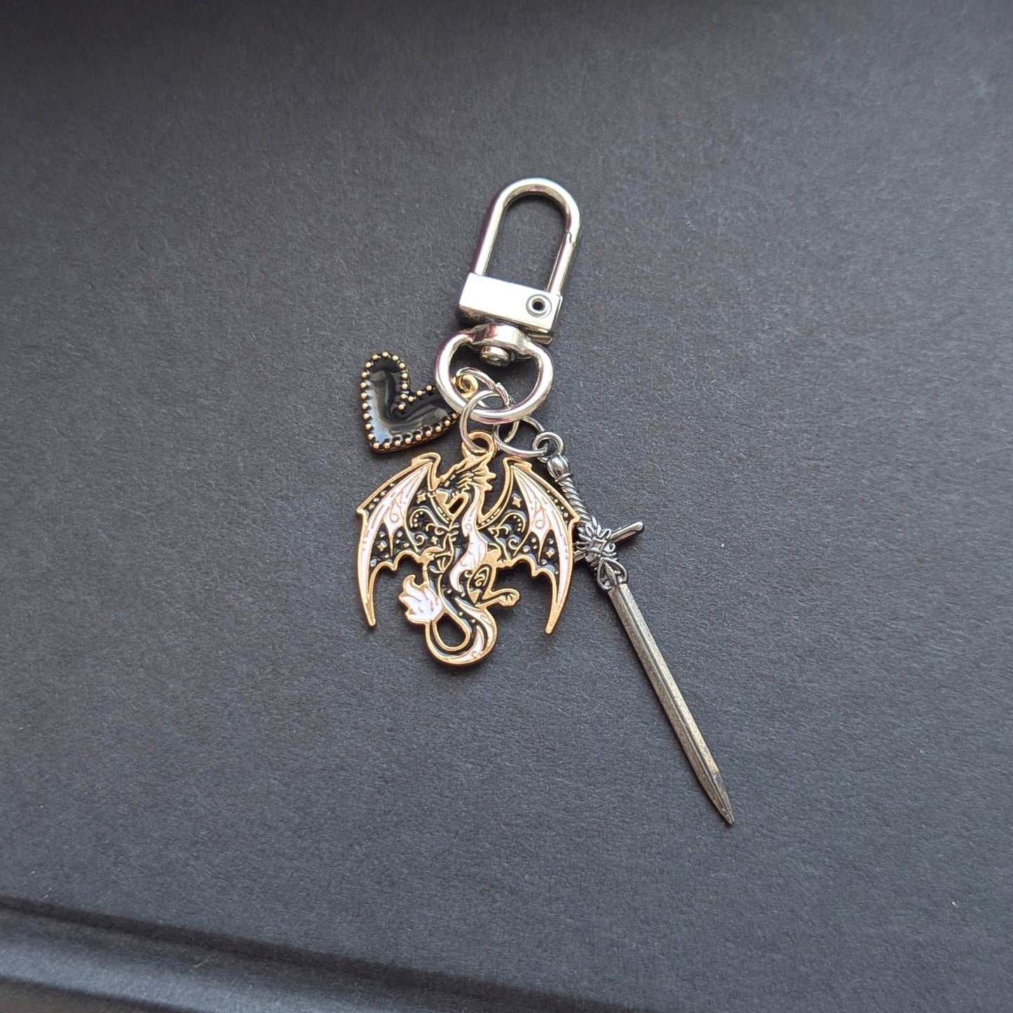 Angie's Creations hand-assembled charm keychain featuring a gold and silver detailed dragon charm with spread wings, a long silver sword charm, and a black beaded heart accent charm on a silver swivel clasp. Fourth Wing and Empyrean series inspired bookish keychain handcrafted in Ottawa Canada.