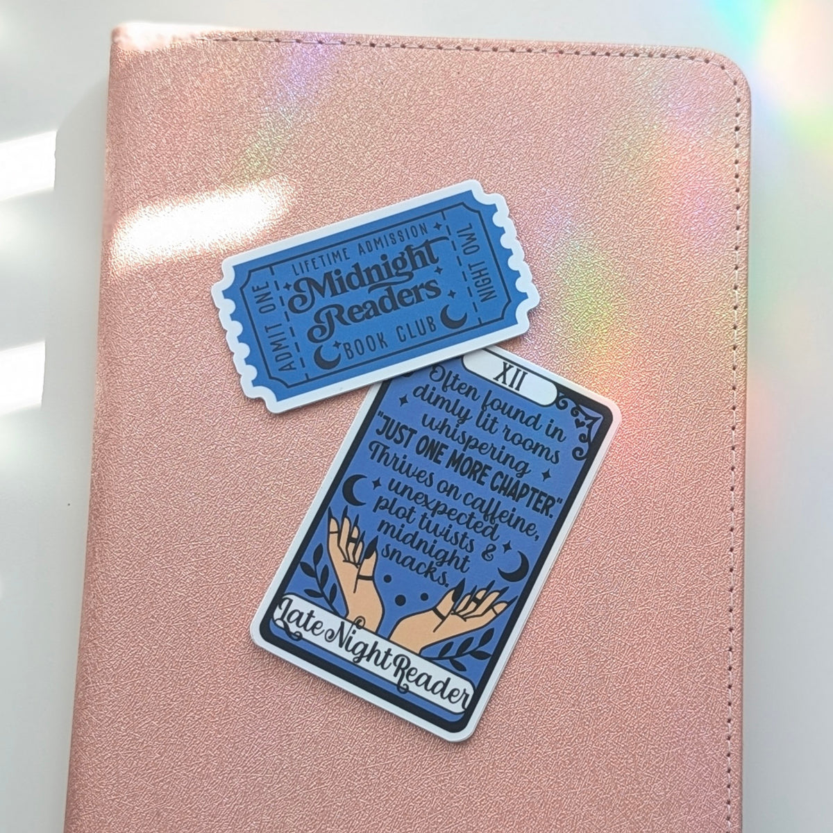 Two decorative stickers featuring a blue and white tarot card design and a blue ticket sticker, placed on a pink iridescent notebook cover.