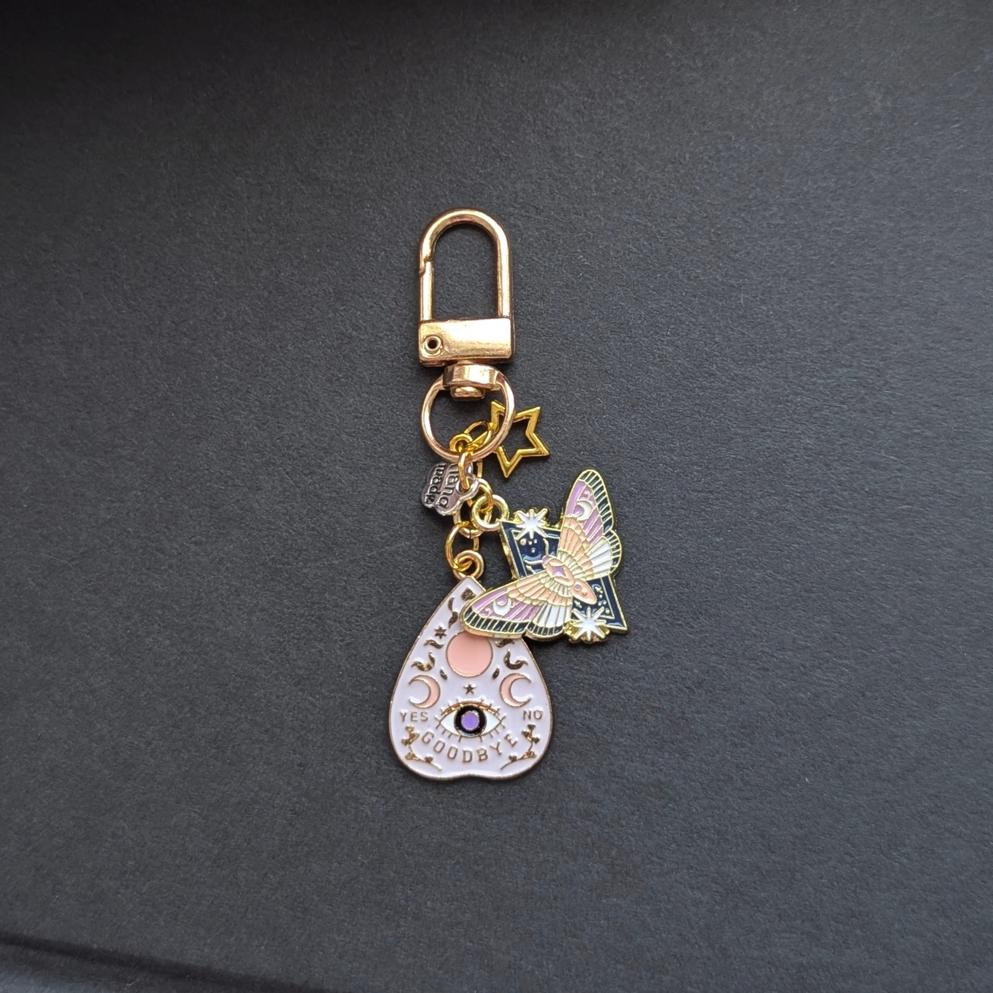 Angie's Creations hand-assembled charm keychain featuring a white and gold enamel ouija planchette charm with moon phase and evil eye detail, a holographic celestial moth charm, a gold star charm, and a small handmade tag, all on a gold swivel clasp. Witchy dark academia romantasy bookish keychain handcrafted in Ottawa Canada.