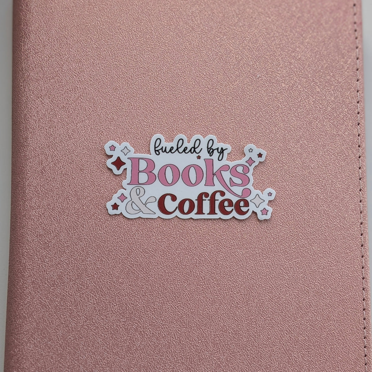 A close-up of a rose gold textured notebook cover featuring a die-cut sticker that reads 'fueled by Books & Coffee' in pink, brown, and black lettering.