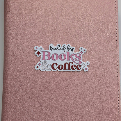 A close-up of a rose gold textured notebook cover featuring a die-cut sticker that reads 'fueled by Books & Coffee' in pink, brown, and black lettering.
