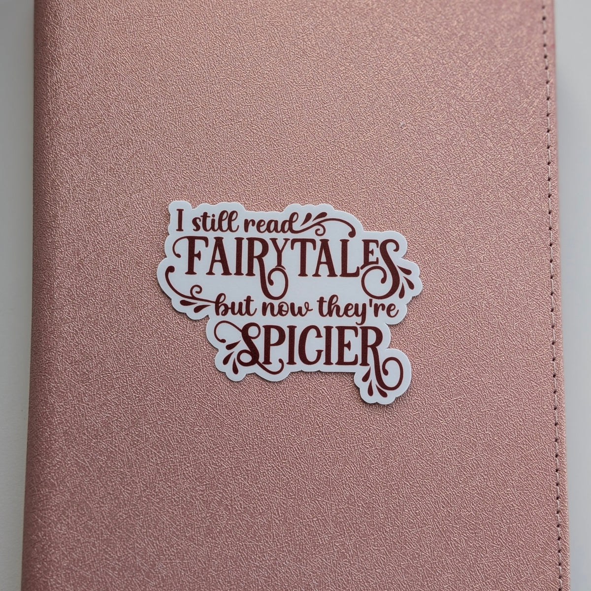 A rose gold faux-leather journal cover featuring a die-cut sticker that reads 'I still read FAIRYTALES but now they're SPICIER' in dark red and white lettering.