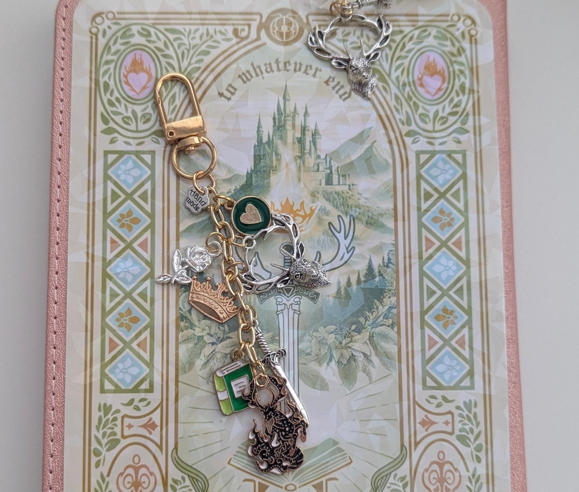 A fantasy-themed keychain charm set displayed on a decorative card. The keychain features a gold lobster clasp, a silver key-shaped charm with a crown, a green heart-shaped charm, a small green gem charm, and a dark metallic key charm, all resting on a pink and cream illustrated card.