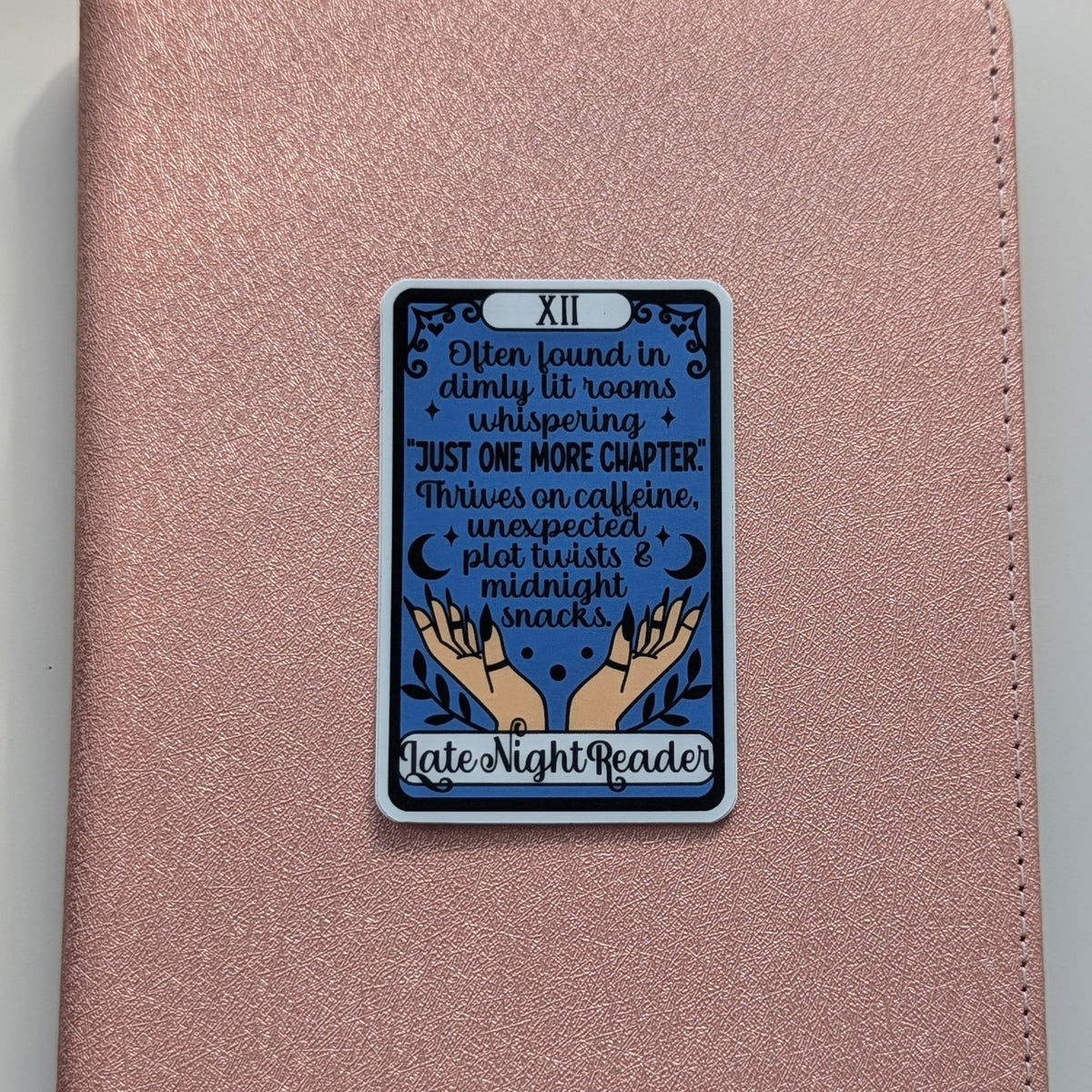 A rose gold textured journal or notebook with a rectangular tarot card sticker on the front. The sticker is labeled 'XII' and features a dark blue background with text reading 'I'm found in dimly lit rooms + whispering + JUST ONE MORE CHAPTER! 