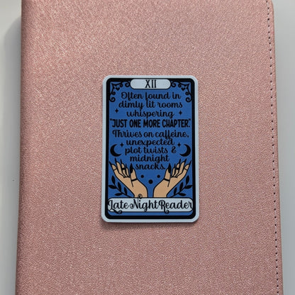 A rose gold textured journal or notebook with a rectangular tarot card sticker on the front. The sticker is labeled 'XII' and features a dark blue background with text reading 'I'm found in dimly lit rooms + whispering + JUST ONE MORE CHAPTER! 