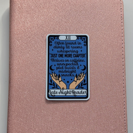 A rose gold textured journal or notebook with a rectangular tarot card sticker on the front. The sticker is labeled 'XII' and features a dark blue background with text reading 'I'm found in dimly lit rooms + whispering + JUST ONE MORE CHAPTER! 