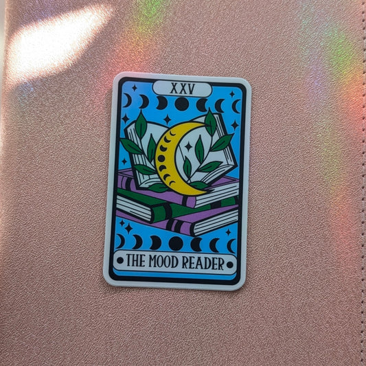 A tarot card sticker featuring 'The Mood Reader' design with a yellow crescent moon, green leaves, and stacked books on a light blue background, placed on a pink textured leather surface.