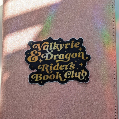 A close-up of a metallic rose gold notebook or planner cover featuring a die-cut sticker that reads 'Valkyrie & Dragon Riders Book Club' in lettering with a black background. The surface is illuminated by warm sunlight with soft shadows and iridescent rainbow reflections.