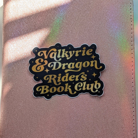 A close-up of a metallic rose gold notebook or planner cover featuring a die-cut sticker that reads 'Valkyrie & Dragon Riders Book Club' in lettering with a black background. The surface is illuminated by warm sunlight with soft shadows and iridescent rainbow reflections.