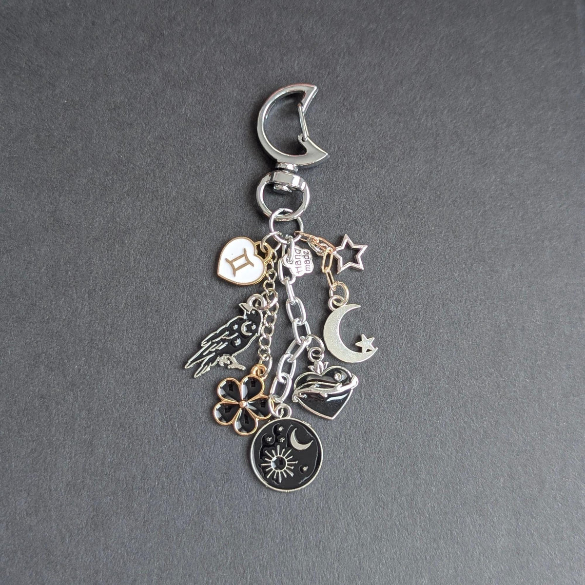 May include: A silver-toned metal keyring featuring a crescent moon clasp and multiple dangling charms, including a white Gemini zodiac heart, a black raven, a black flower, a sun and moon disc, a black heart, and a small silver star.