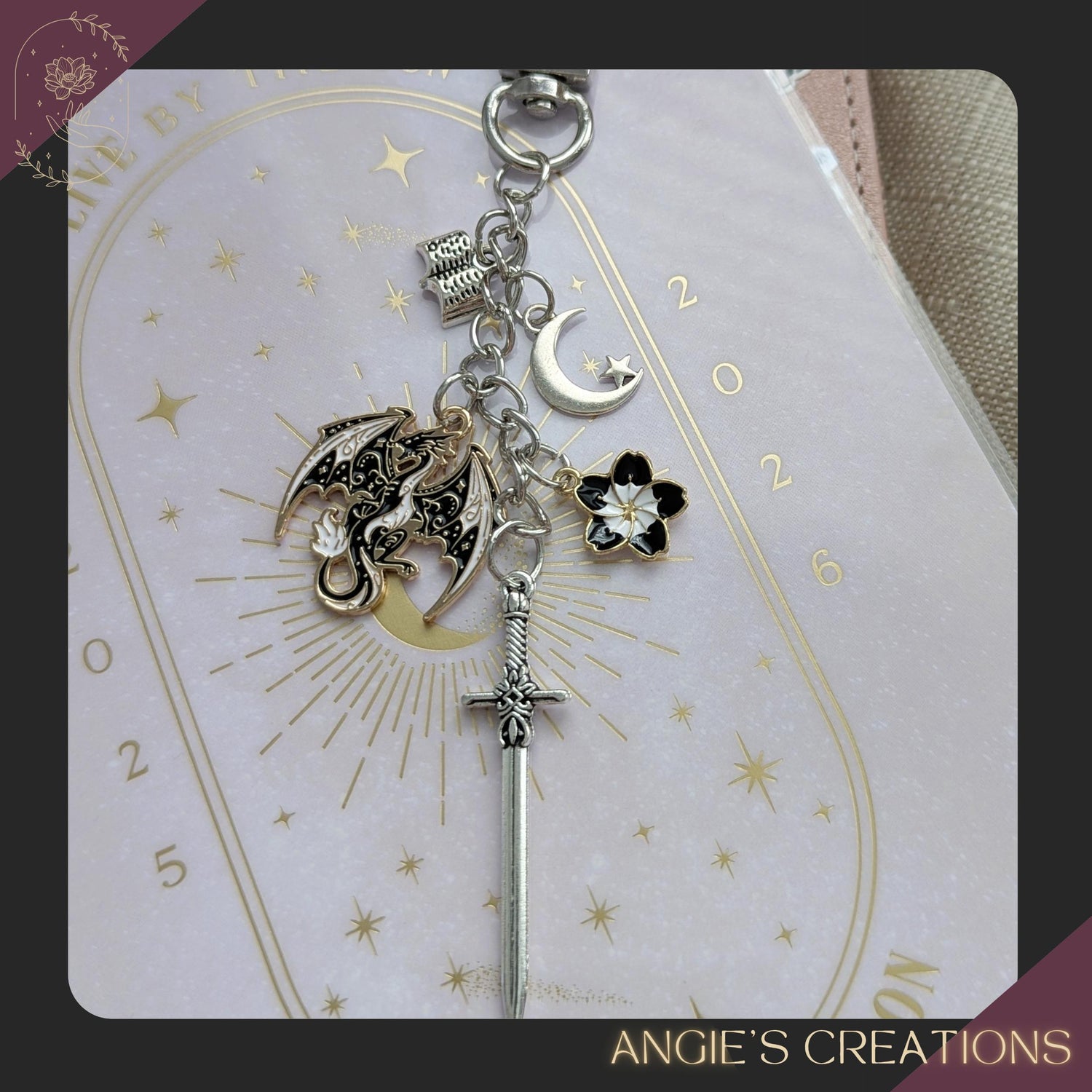 A handmade romantasy-themed Kindle charm chain featuring a detailed dragon charm, silver sword, crescent moon, flower charm, and tiny open book, attached to a silver lobster clasp. Perfect for fantasy readers and bookish accessory lovers.