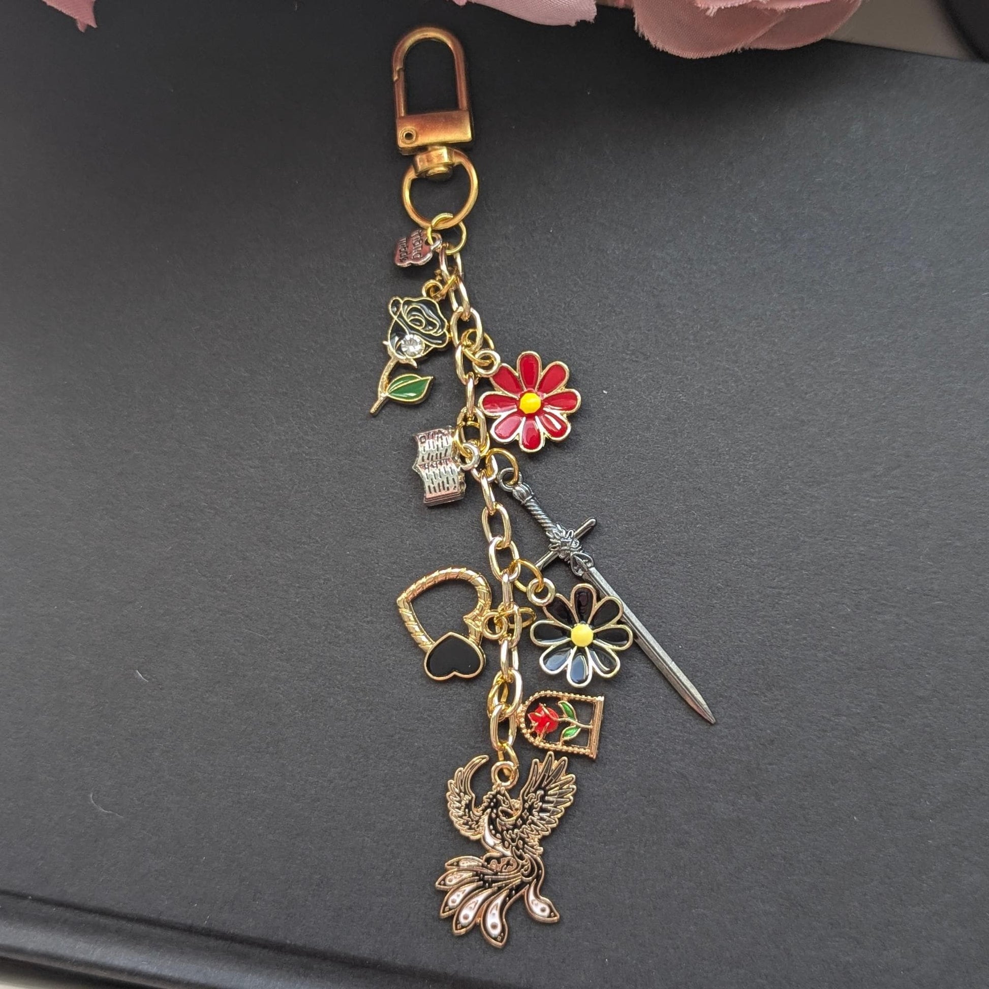 May include: Gold-coloured keychain with various charms. Charms include a red and yellow flower, a sword, a heart, a phoenix, and a rose. The keychain is attached to a gold clasp.