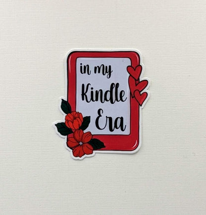 May include: A sticker featuring the phrase "in my Kindle Era" in black script, within a red border. The design includes red hearts and flowers with green leaves, set against a white backdrop.