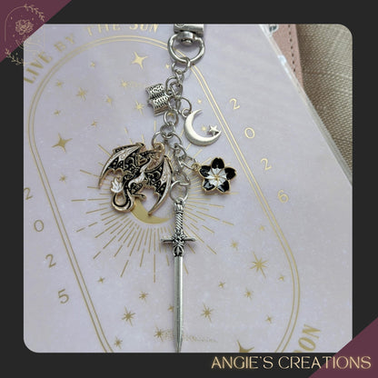 A handmade romantasy-themed Kindle charm chain featuring a detailed dragon charm, silver sword, crescent moon, flower charm, and tiny open book, attached to a silver lobster clasp. Perfect for fantasy readers and bookish accessory lovers.
