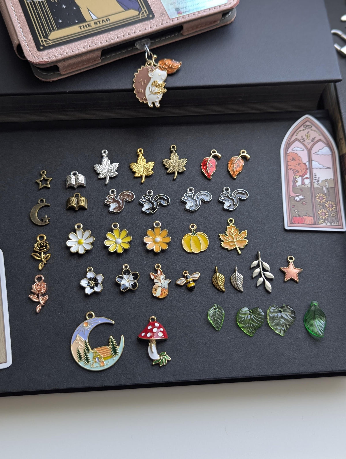 May include: A collection of assorted metal and enamel charms featuring various shapes including squirrels, leaves, flowers, a pumpkin, a bee, a mushroom, a crescent moon with a landscape, stars, and books, arranged on a dark surface.