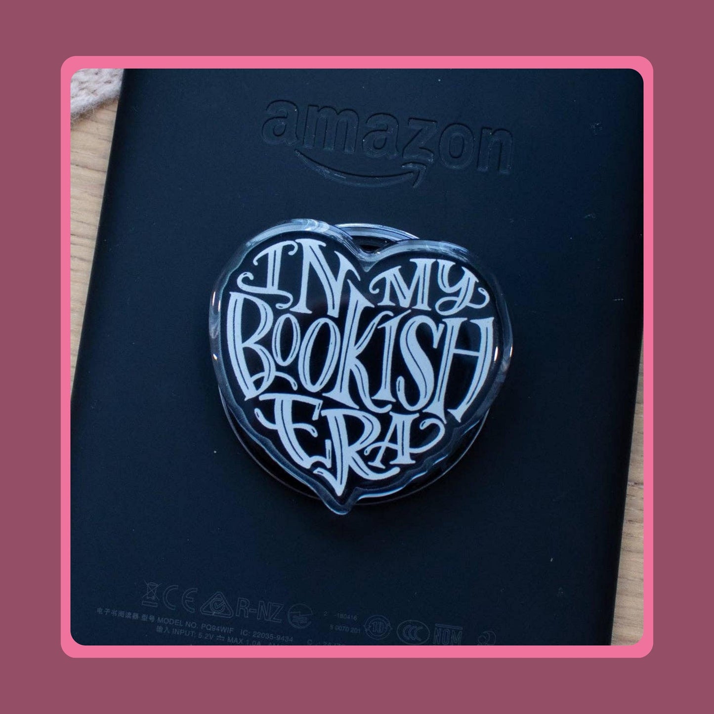 Black heart-shaped acrylic charm with white "In My Bookish Era" script lettering, sold as a matching Kindle grip and phone dust plug set.