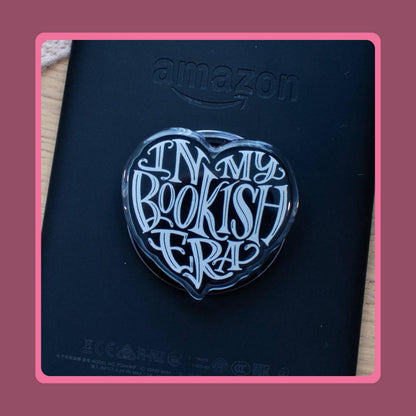 Black heart-shaped acrylic charm with white "In My Bookish Era" script lettering, sold as a matching Kindle grip and phone dust plug set.