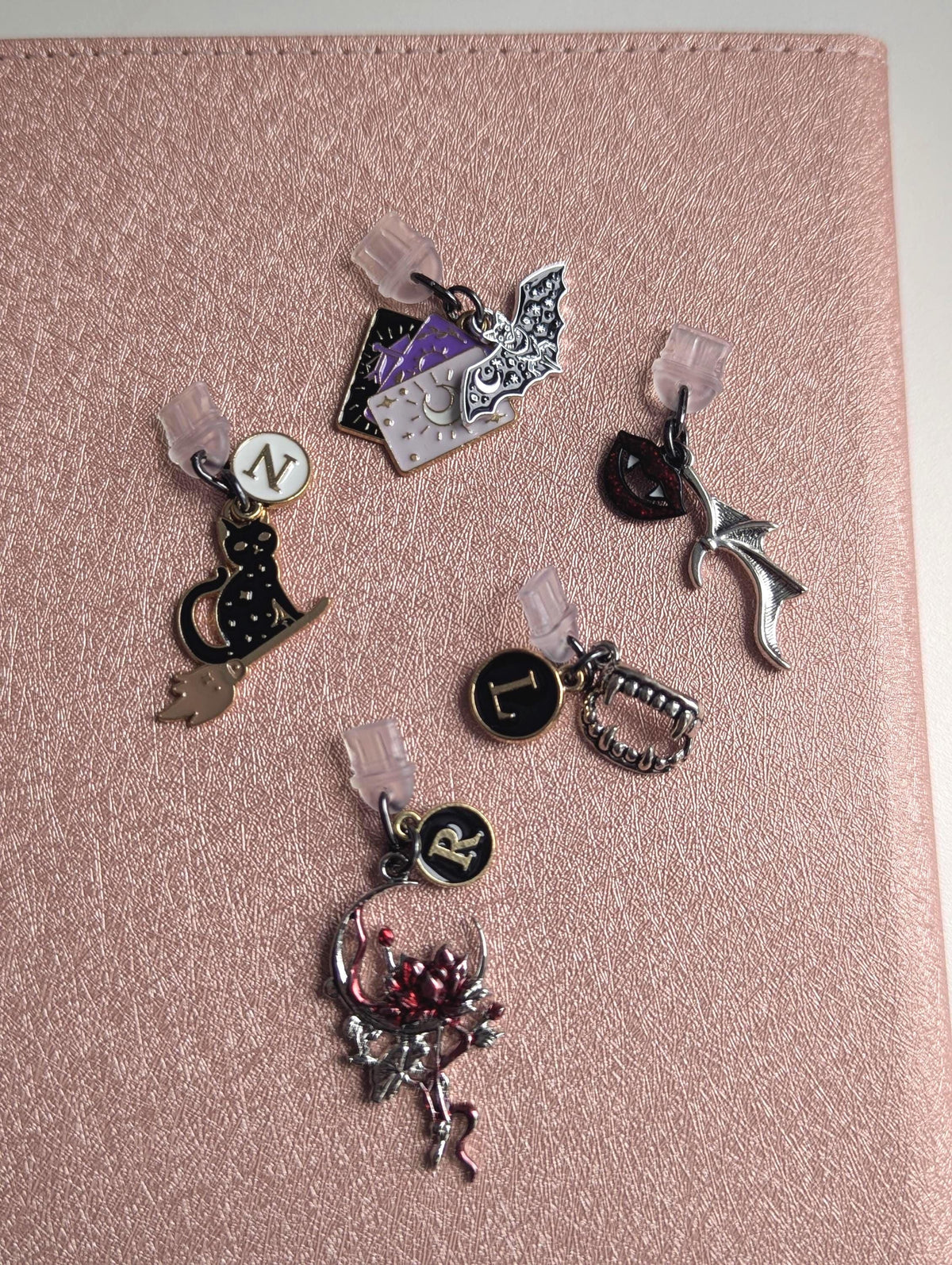 May include: Five metal charms with clear plastic attachments. One features a black cat on a broom with the letter N, one a bat and tarot cards, one a red lip and wing, one a vampire fang with the letter L, and one a red flower with the letter R.