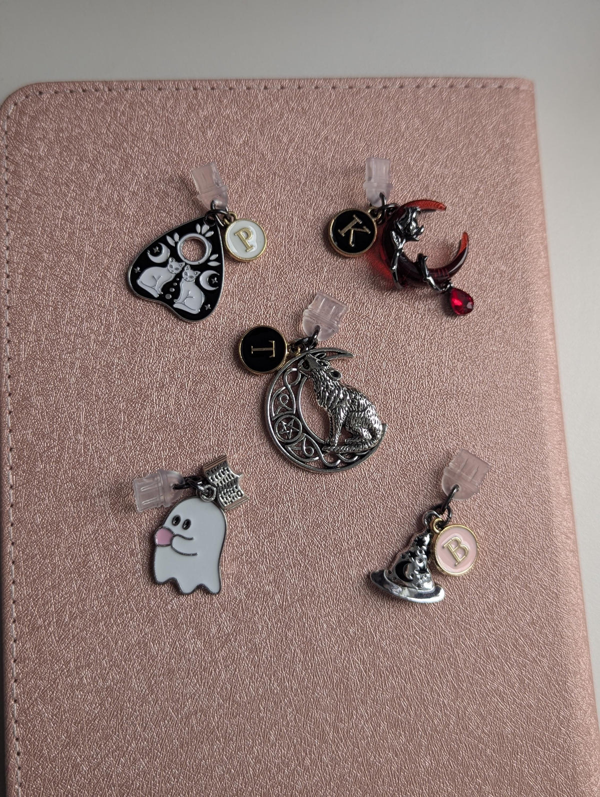 May include: Five metal charms with clear plastic attachments. One is a black planchette with white cats, one a red crescent moon with a cat and letter K, one a silver wolf on a moon with letter T, one a white ghost, and one a hat with letter B.