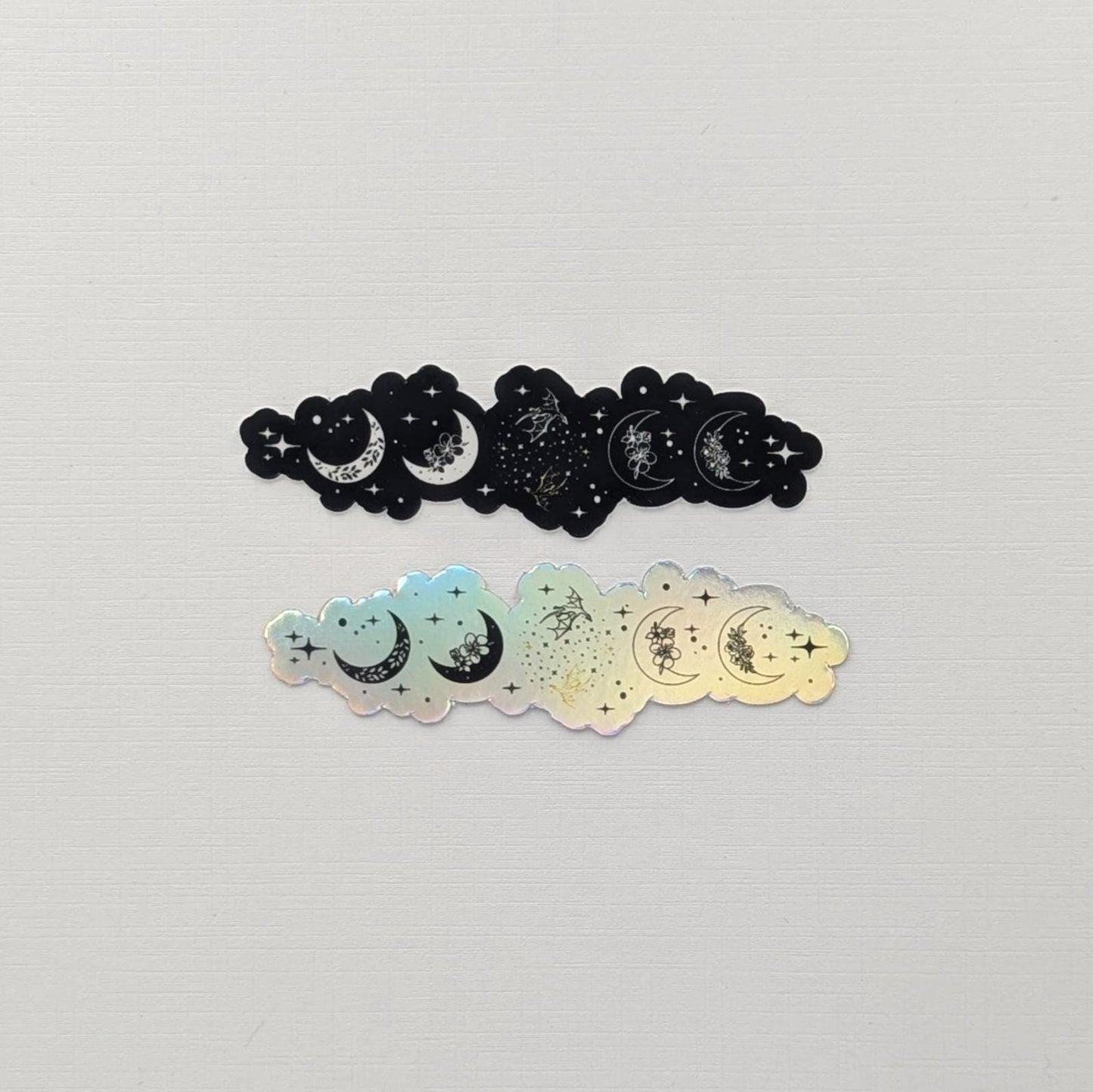 May include: Two stickers with a celestial design. One is black, the other is iridescent. Each sticker has a cloud shape with crescent moons, stars, and floral details. Ideal for crafting and DIY projects, these stickers add a touch of magic.