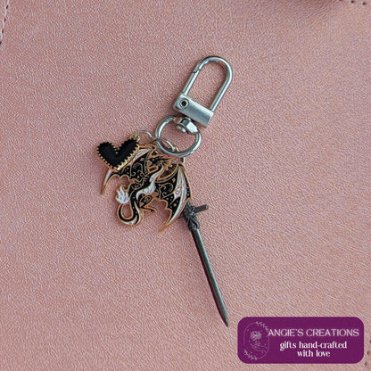 May include: A keychain featuring a dragon charm, a black heart charm, and a small sword charm. The dragon charm is black, white, and gold. The keychain has a silver clasp. The text "ANGIE'S CREATIONS gifts hand-crafted with love" is on a purple label.